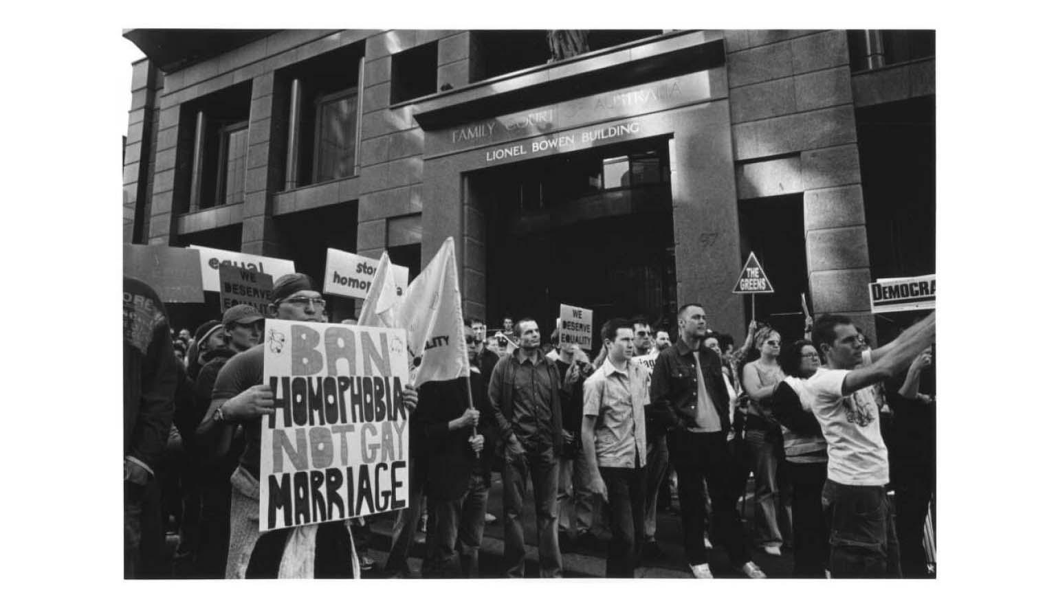 A black and white photo of a large number of people marching through city streets with signs supporting gay marriage. One sign says 'Ban homophobia, not gay marriage'.
