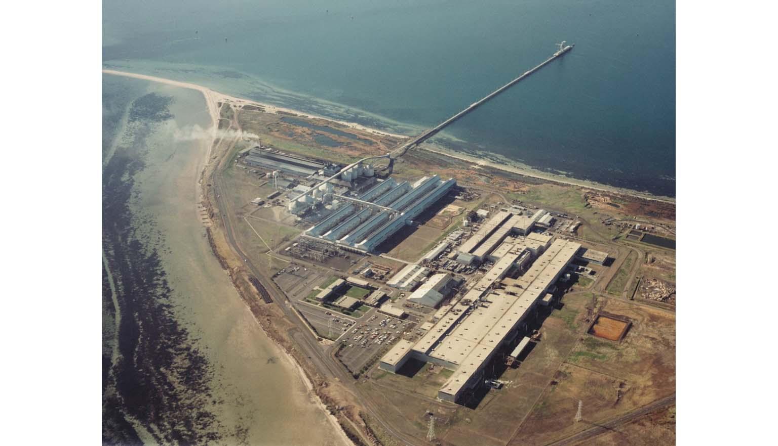 An aerial shot of a huge factory on a point surrounded by ocean. The factory is made up of several large sheds and warehouses. A long pier runs out into deep water.