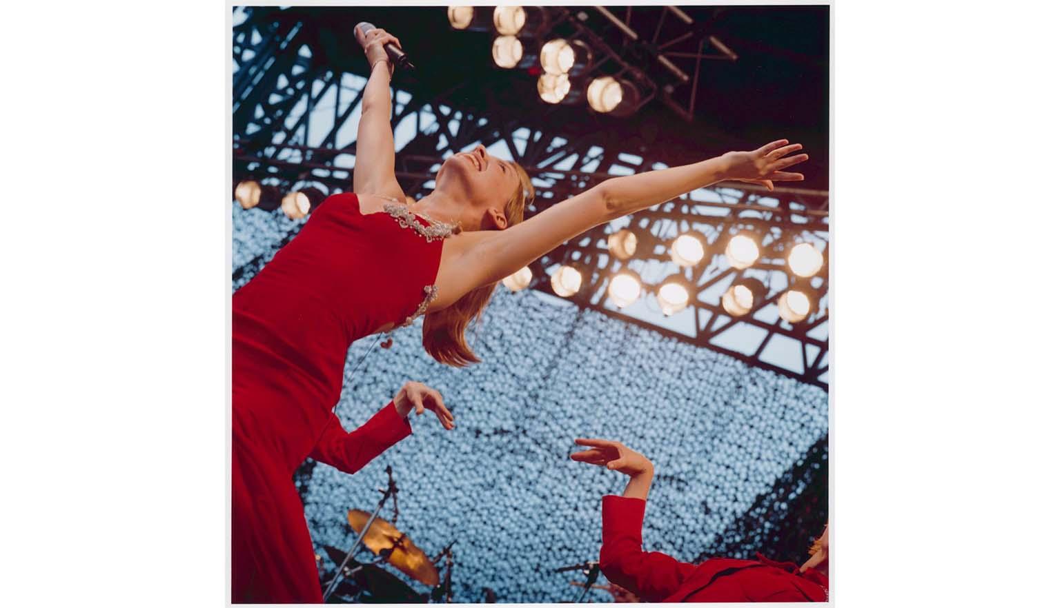 A woman in a red dress throws her hands up in the air while on stage. Behind her a backup dancer is performing. Many spotlights are shining on the stage