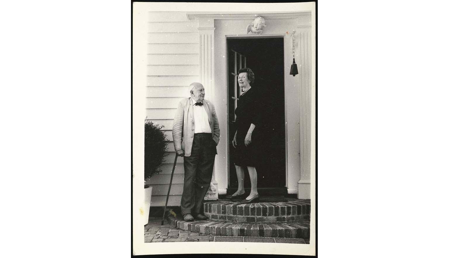 A black and white photo of an old balding man in a black bowtie, leaning on his cane and a woman in a black dress, smiling, about to enter a large white portal doorway, with a large black ship's bell hanging next to it.
