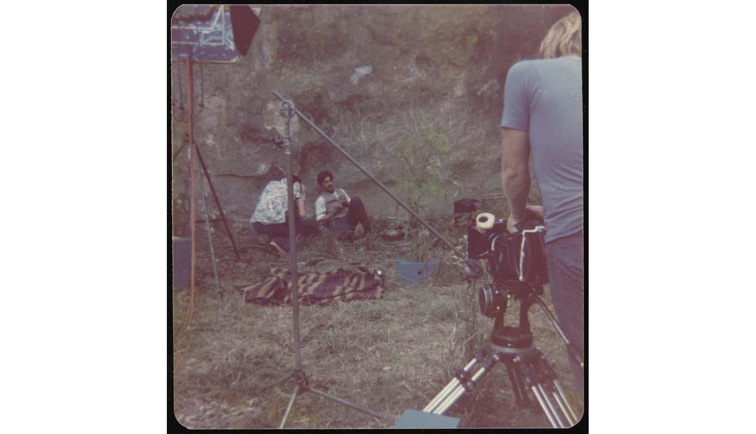A man with filming equipment filming two actors at base of mountain