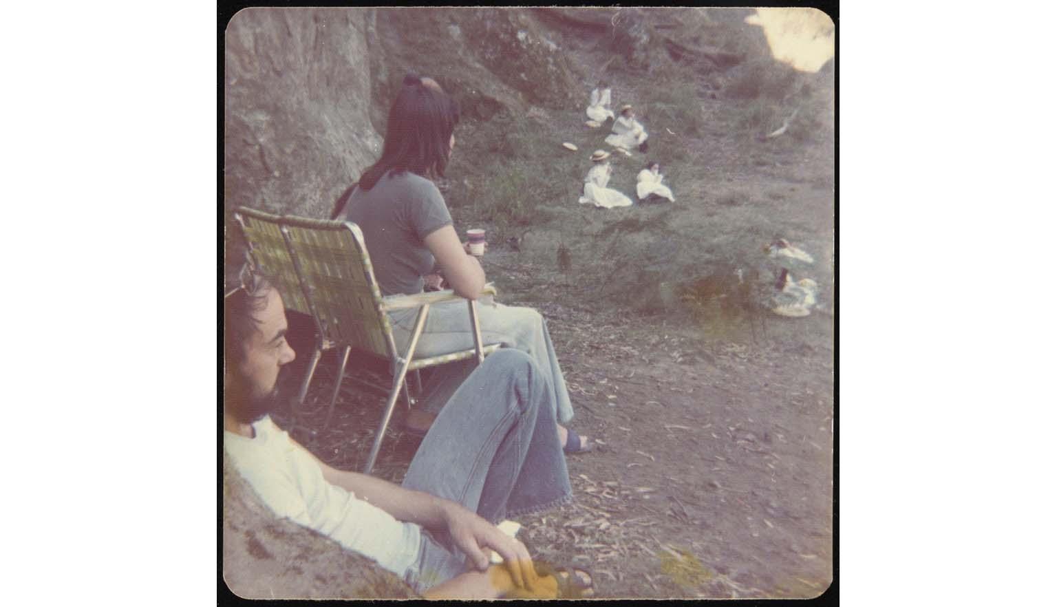 Three people in beach chairs watch filming group of girls in white Georgian dress