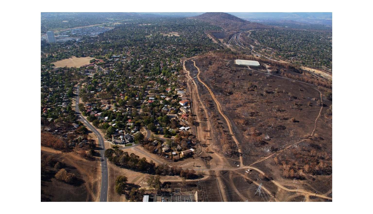 An aerial shot of a city suburb. A hill surrounding the suburb is scarred black from a large fire.