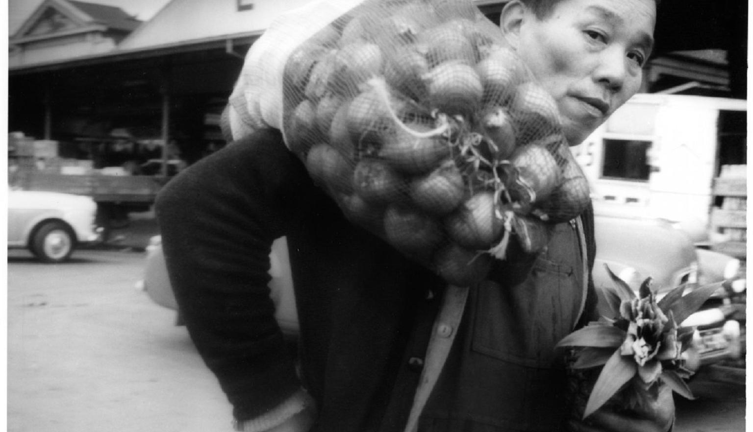 A black-and-white photograph of a man carrying a large sack of produce over his shoulder while holding a small plant. He stands in front of market buildings and cars.