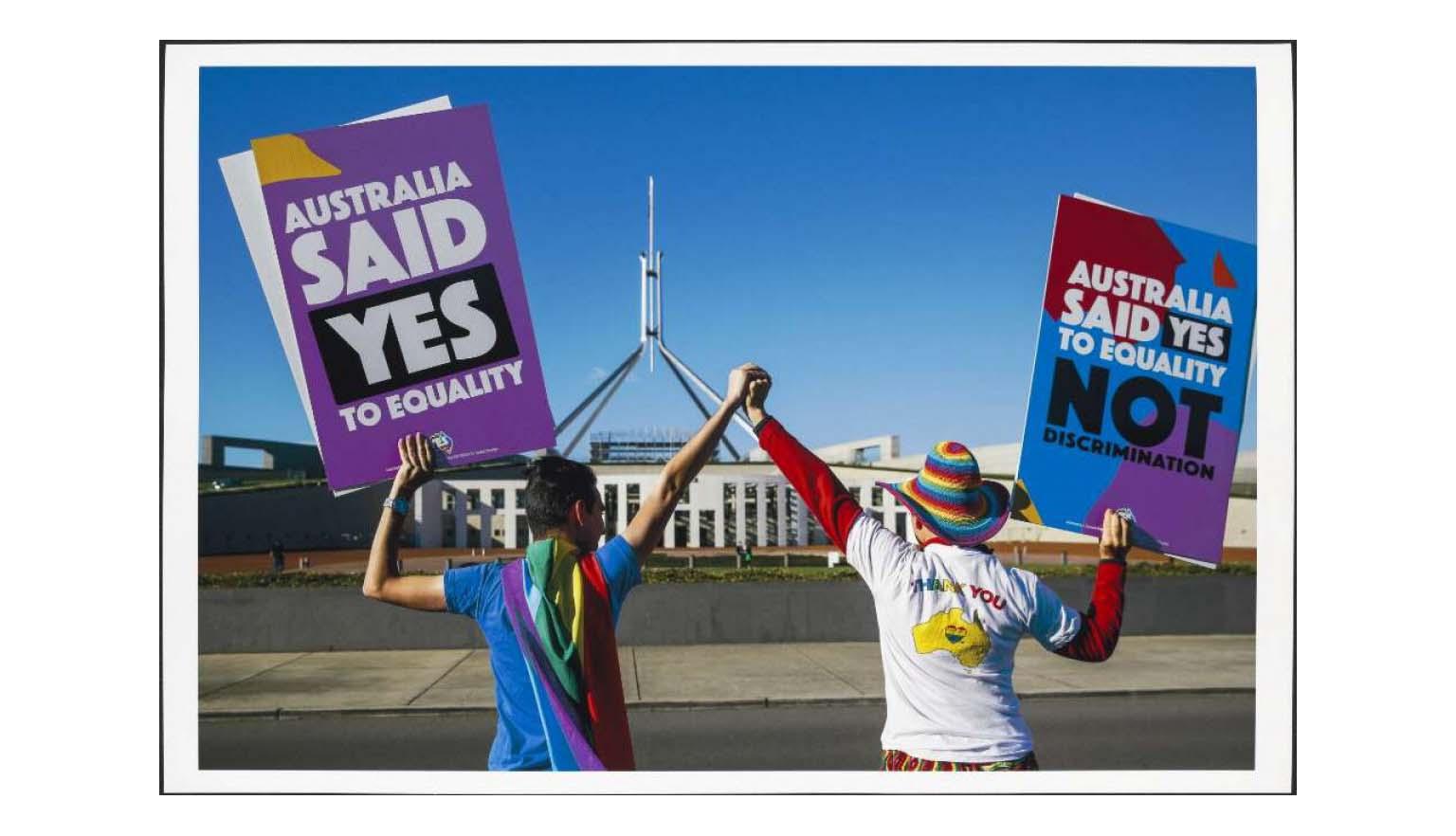 A photo of 2 people with their backs to the camera, facing Australia's Parliament House, holding hands, lifting them high. Each is holding a large sign in their outside hands that reads 'Australia said Yes to equality' and 'Australia said Yes to equality, not discrimination'.