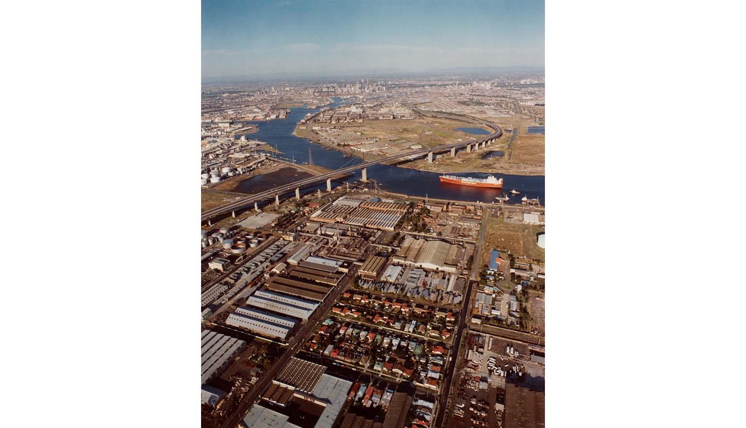 An aerial shot of a city port industrial district. A large river winds through the middle. A large ship is on the river about to sail under a bridge.