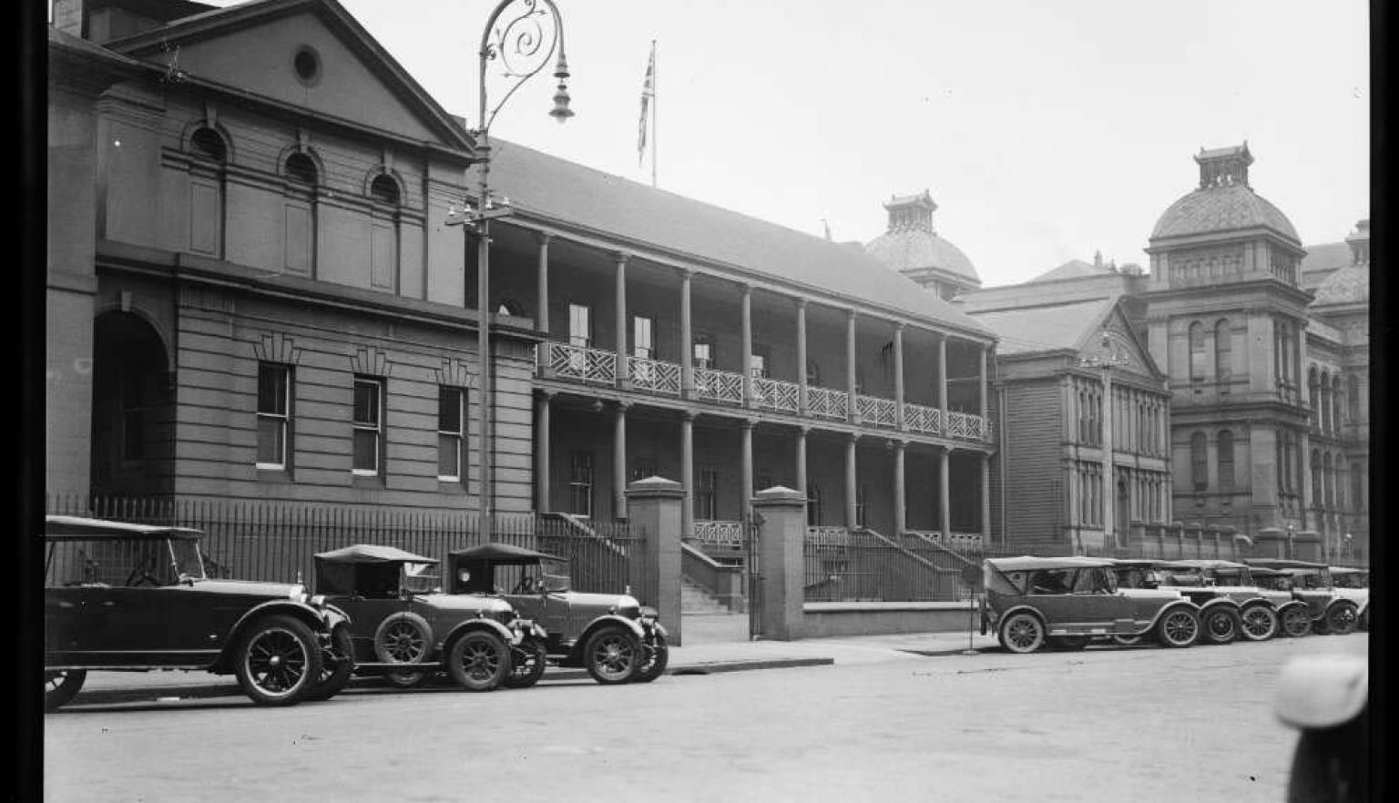 black and white photo of old buildings with old cars in the street out the front