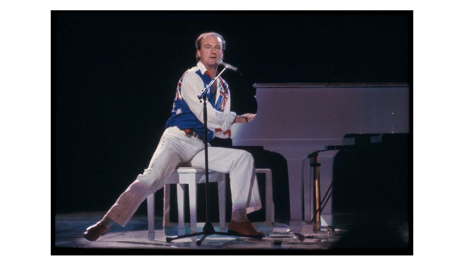 A man wearing white pants, shirt and an Australian flag vest plays a white grand piano on stage