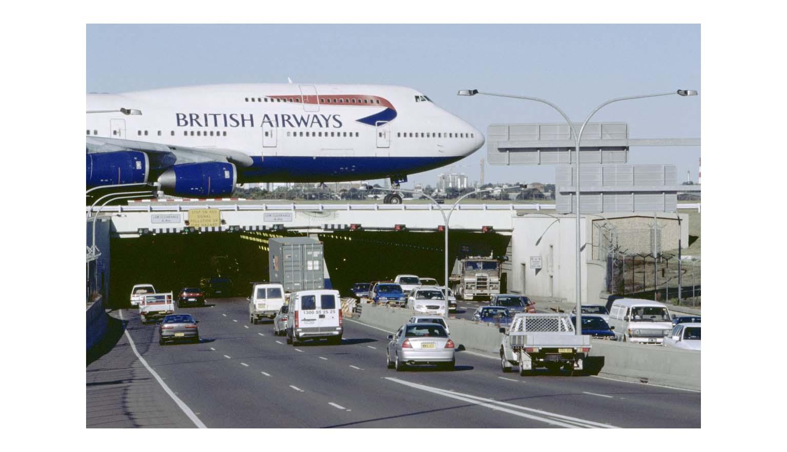 A British Airways Boeing 747 Jumbo Jet taxis across the runway bridge over a highway tunnel. Cars are entering and exiting the tunnel