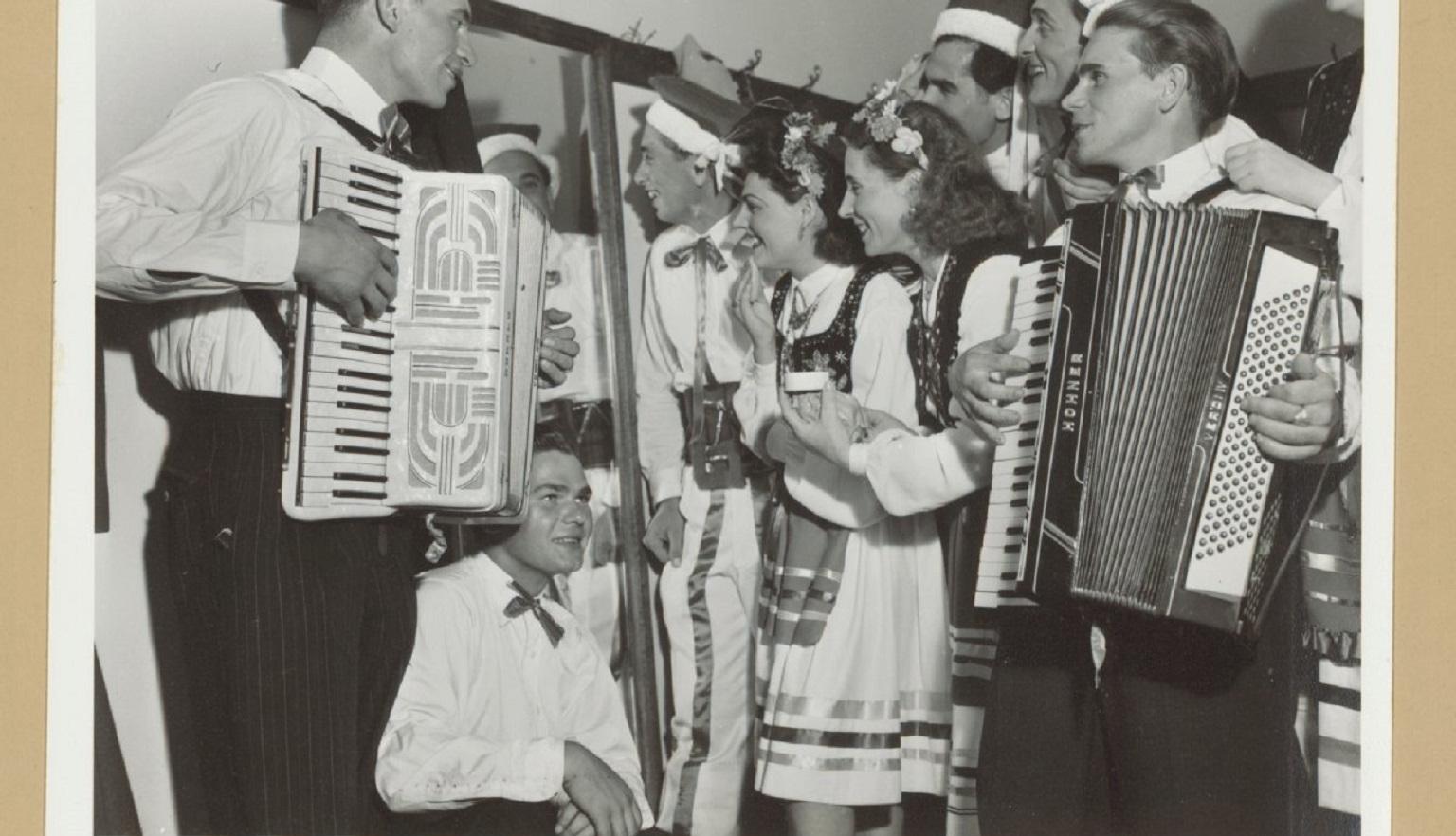 A black-and-white photograph of a group of people in traditional Polish folk costumes. Two men play accordions, while others stand and smile, some in conversation.