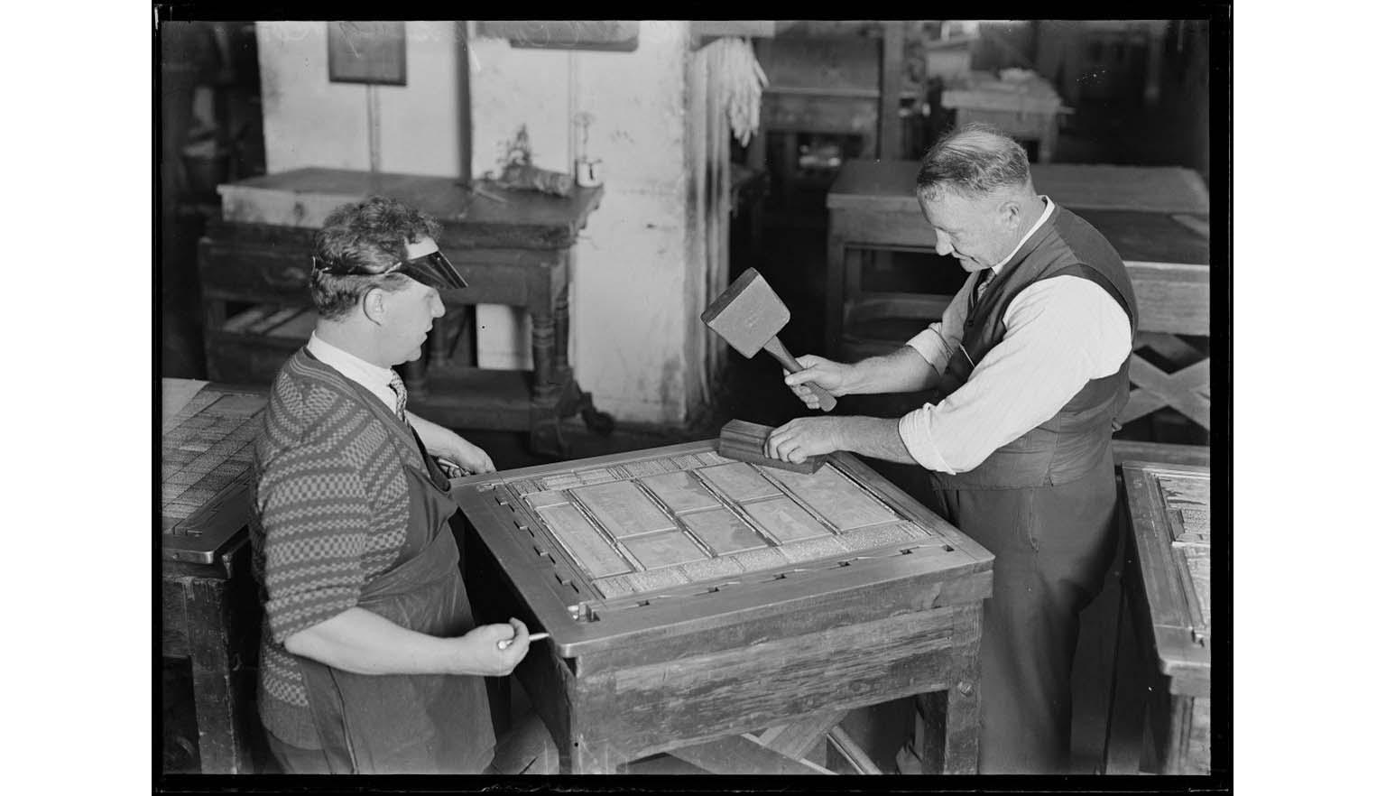 A black and white image of two men setting a page up with typeface. One man is setting the screws of the frame while another man makes  sure all the letters are set by hitting them with a wooden mallet.