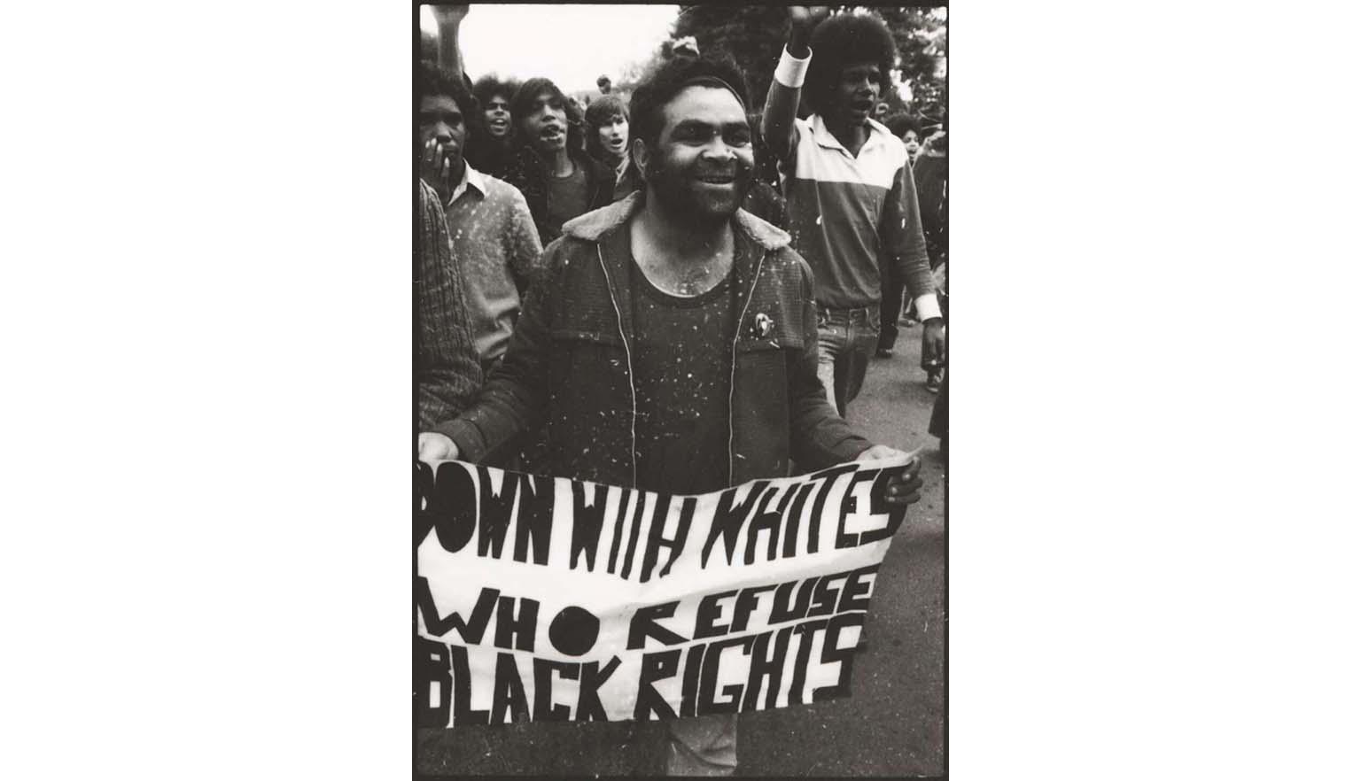 A black and white photo of an Indigenous Australian carrying a sign in a rally of black and white people, that says 'Down with whites who refuse black rights'.