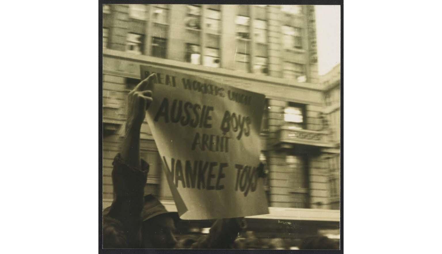 A sepia toned photo of a man in a hat holding a paper sign in a city street, saying 'Meat Workers union Aussie boys aren't Yankee toys'
