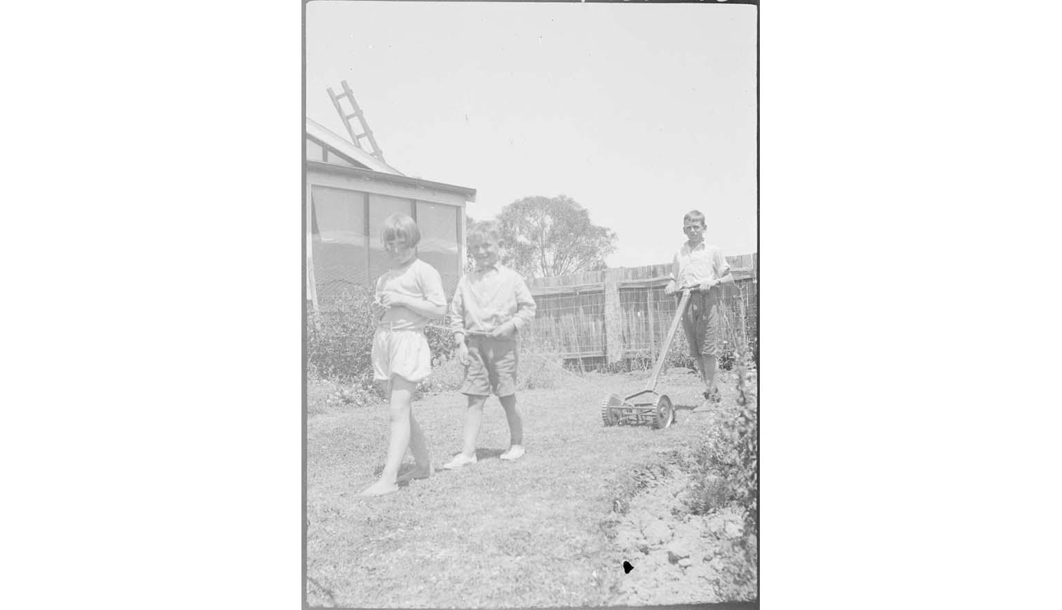 black and white photo of two young boys standing in front of their father mowing the lawn with manual mower