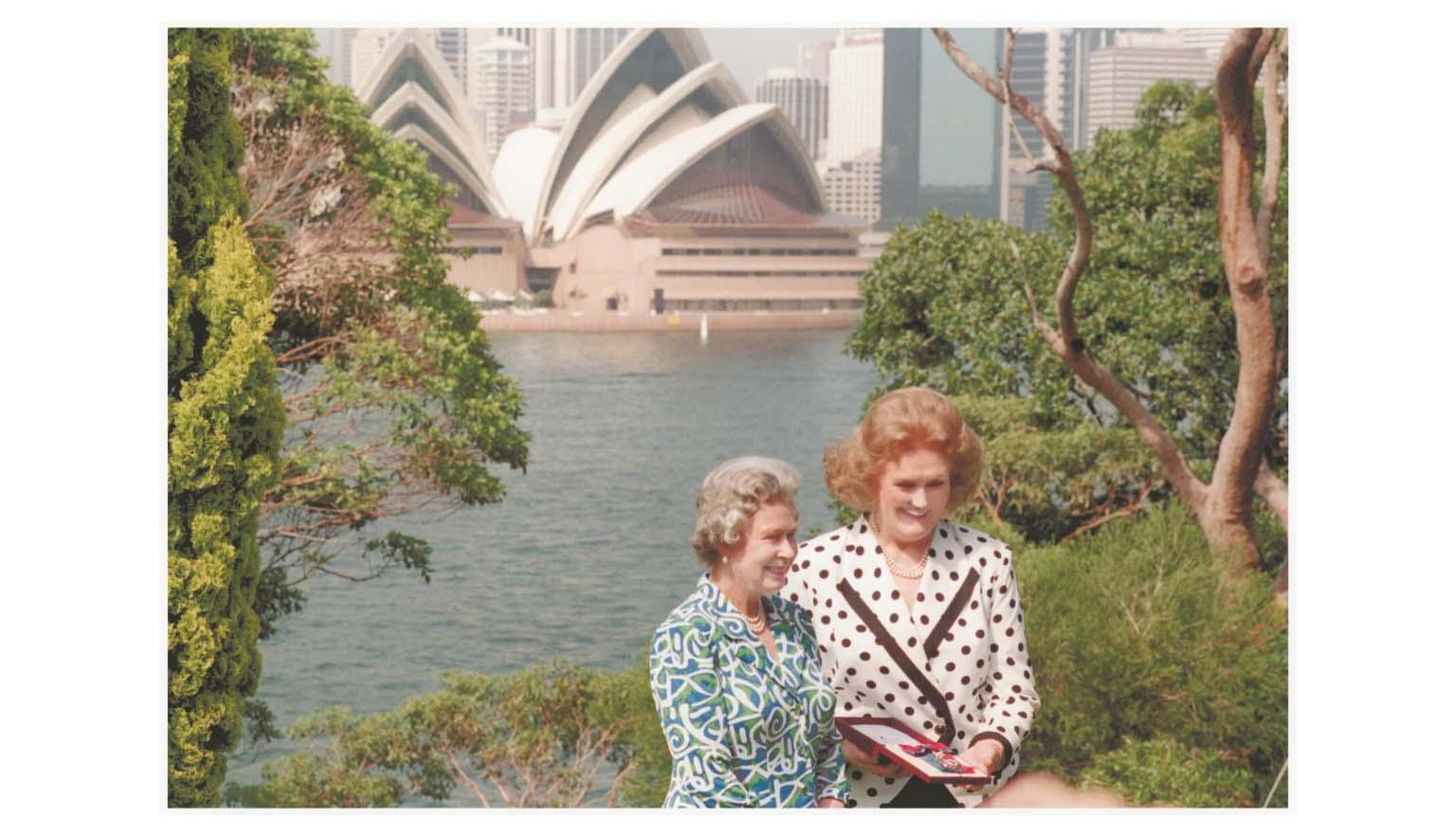 Two women stand among trees with the Sydney Opera House in the background. One lady wears a white and black polka dot blazer and has red hair. The other wears a green, white and blue patterned dress. She has grey hair. Both are smiling