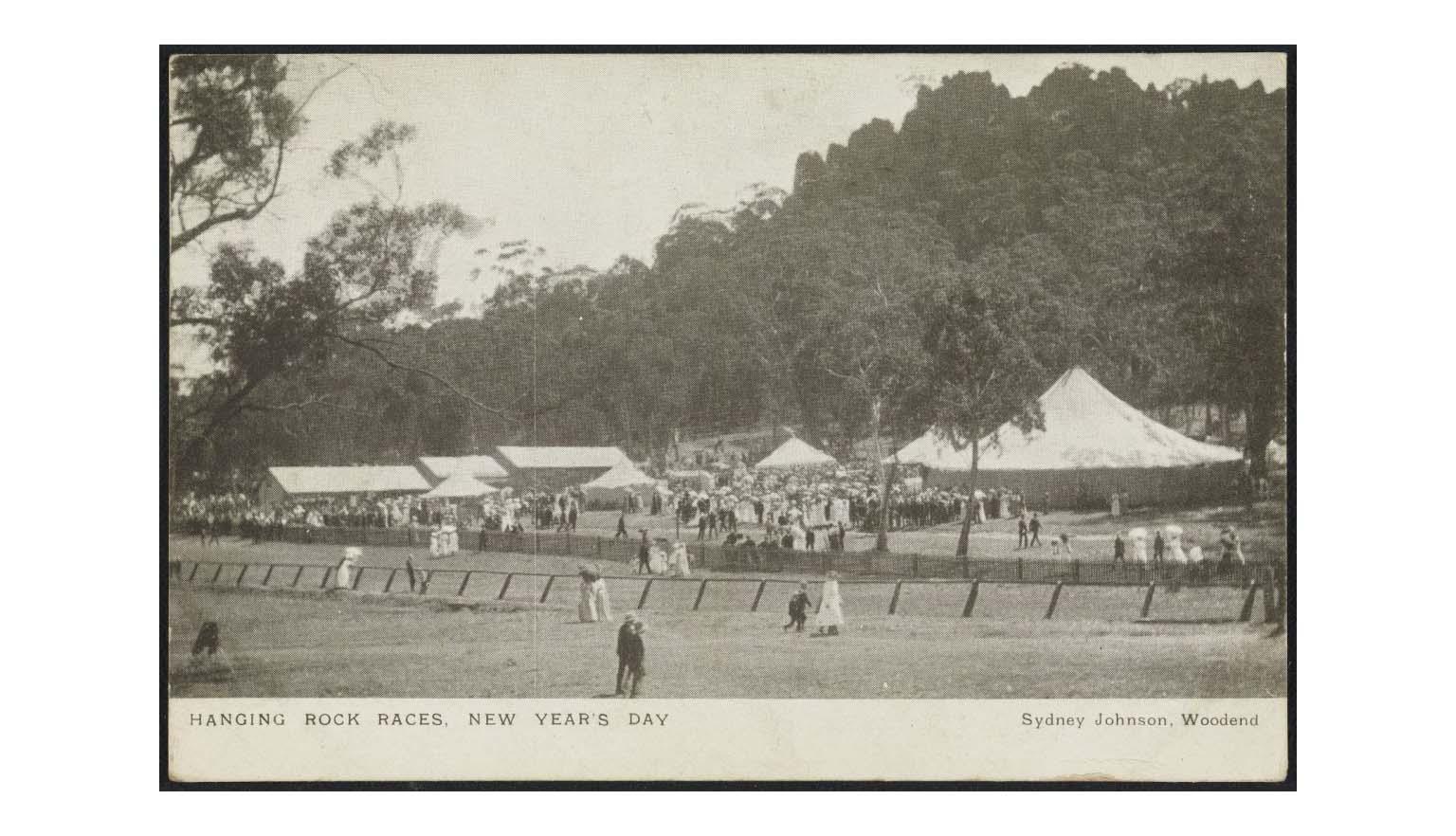 An old black and sepia toned photo of part of a race course, several tents and hundreds of people dressed in clothes from 1910, and a rocky topped mountain. 