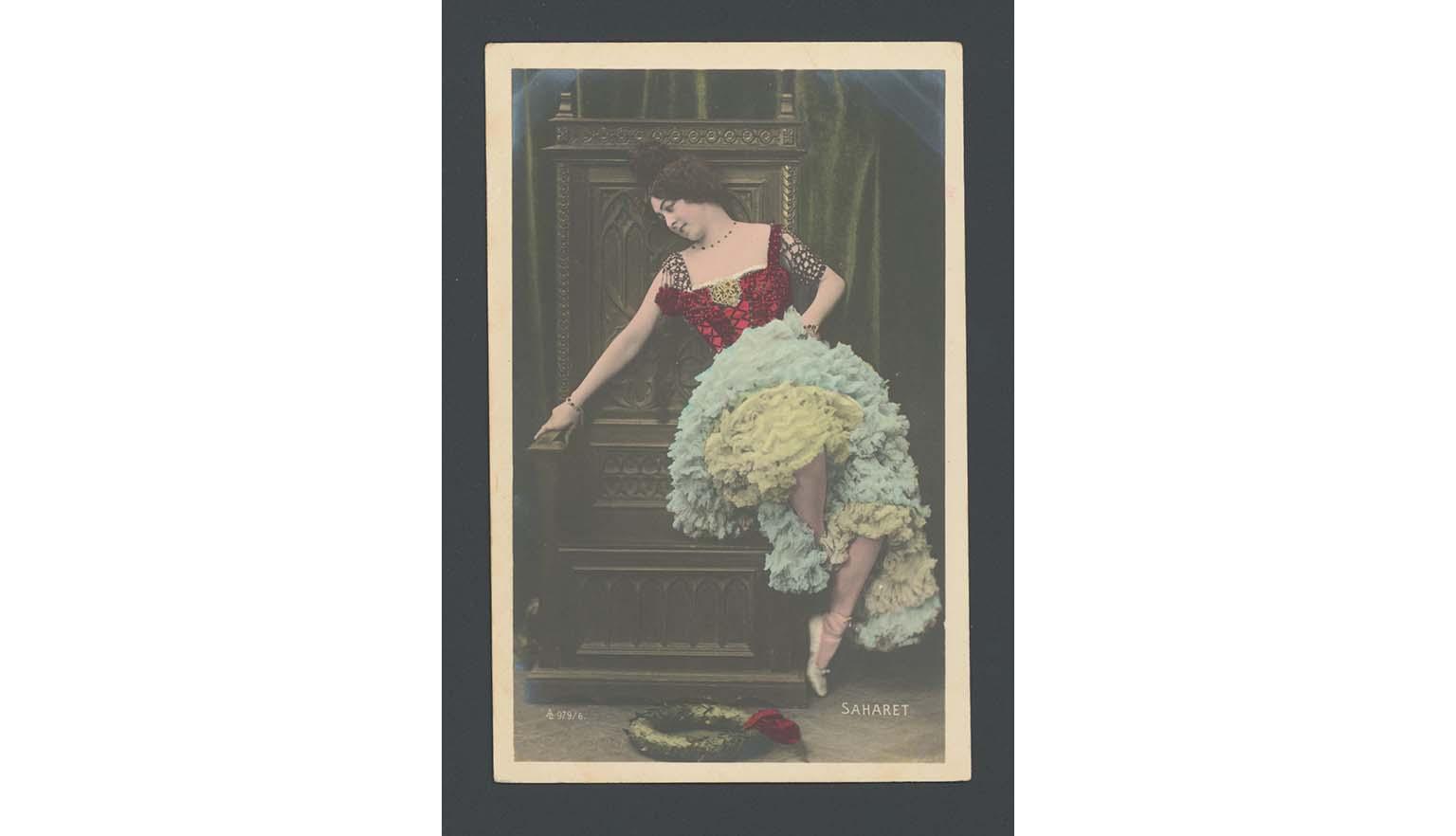 A vintage colourised photograph of a woman sitting on a the arm of a large throne or chair. She is wearing a dress with a red bodice and multiple layers of frills in blues and yellows. She has ballet shoes on. On the floor in a wreath.