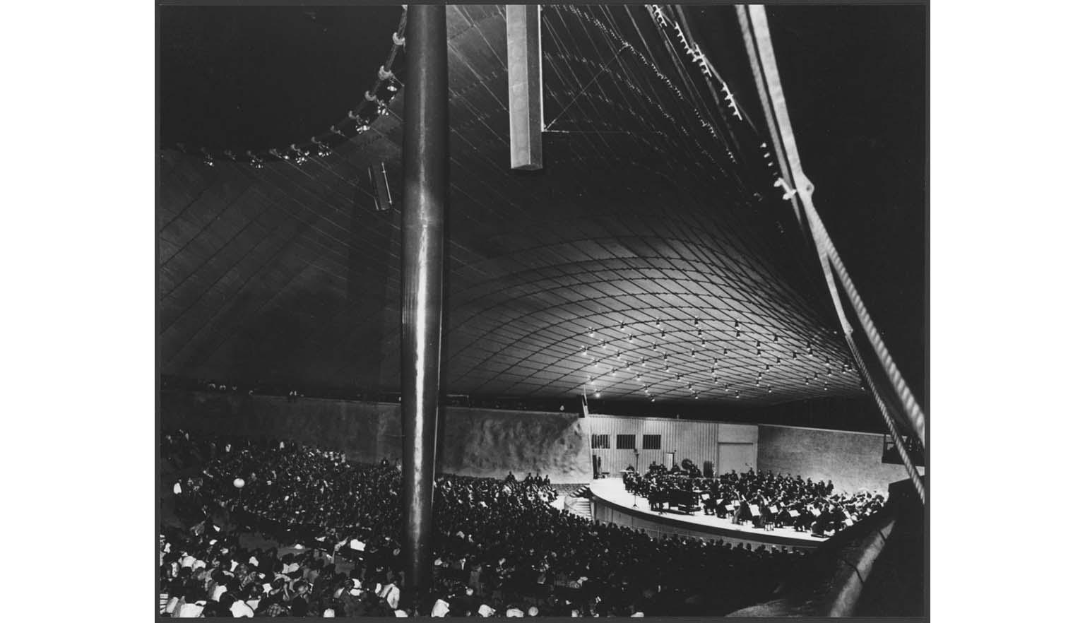 A black and white photograph of the Sidney Myer Music Bowl. A large crowd is watching a performance of an orchestra on the stage.