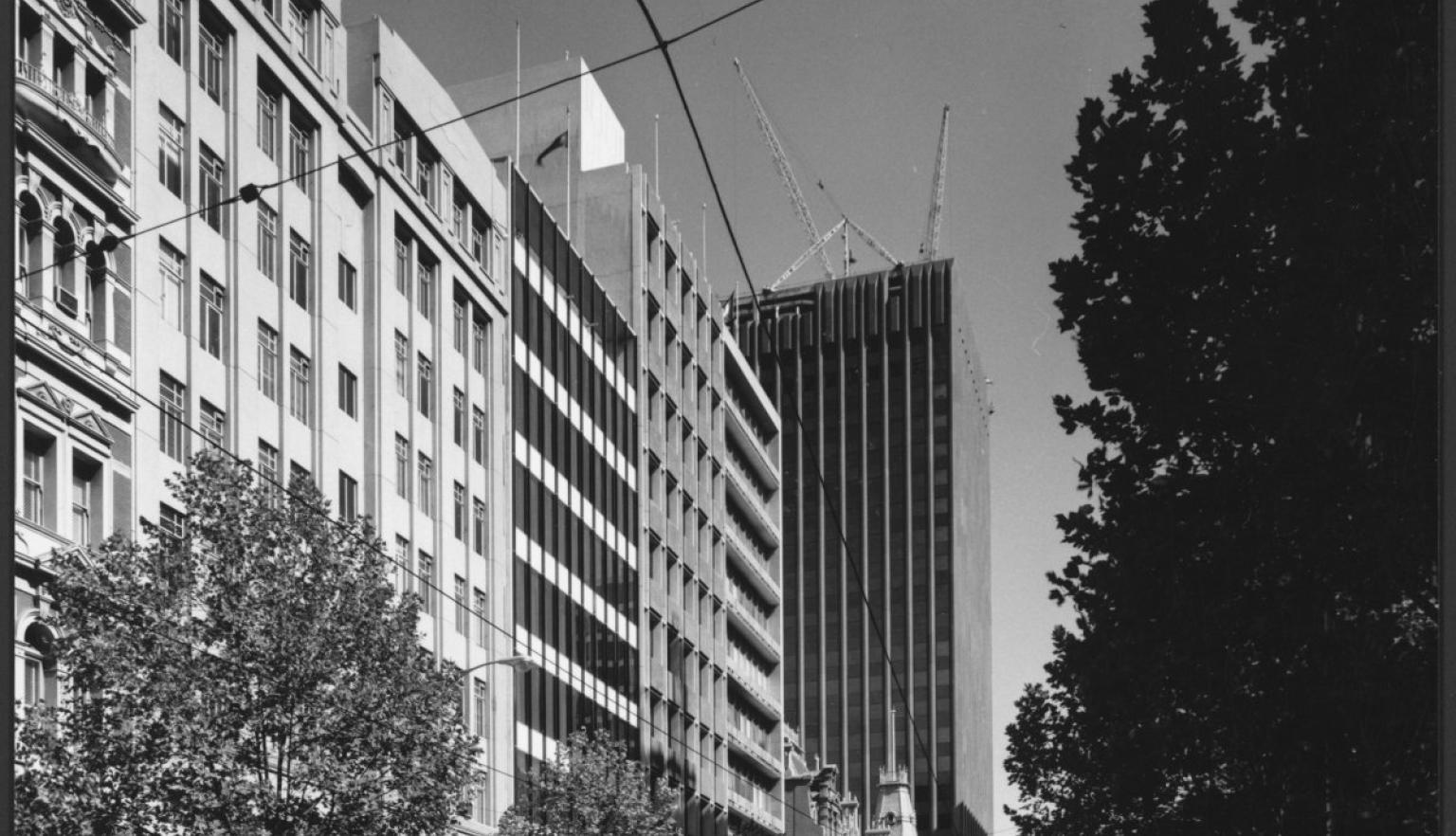 A black-and-white photograph of Bourke Street, Melbourne, showing mid-century modern buildings, with the AMP-St. James building under construction. Cars are parked along the street, with trees lining the road.