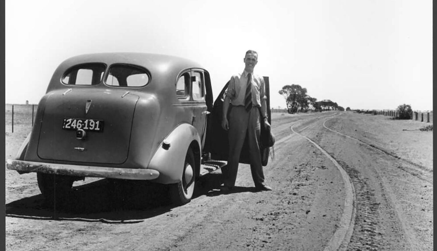Black and white image of a man in a suit standing next to a car on a dirt road. 