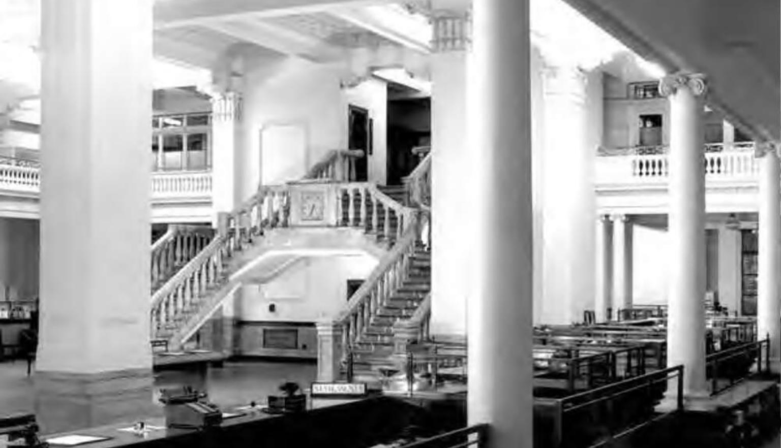 Black and white photo from inside the National Bank head office. 2 ornate staircases in the middle of the image surrounded by white pillars and desks. 