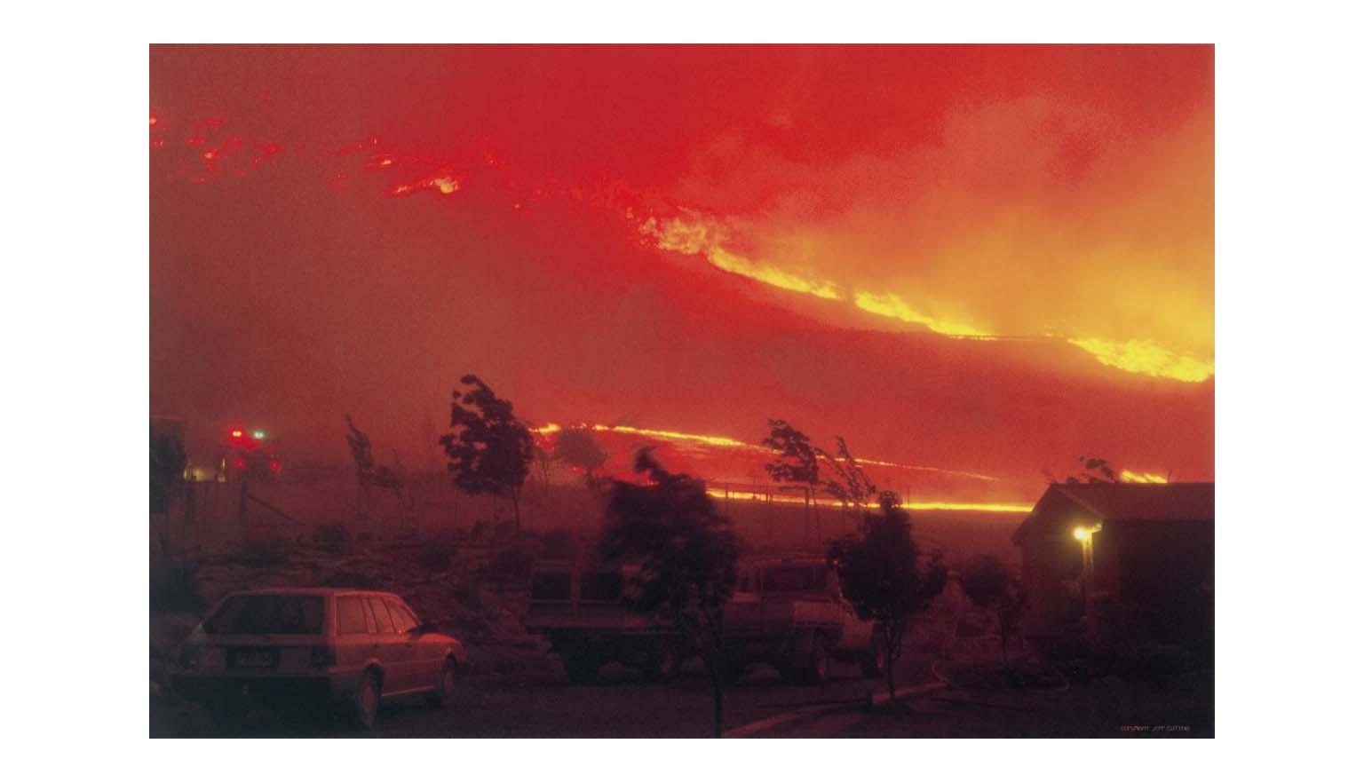 A scene of a hillside completely ablaze. A large fire front is moving up the hillside. The image is blanketed with smoke. The lights of a fire engine can be seen through the smoke. A station wagon is parked in the forground.
