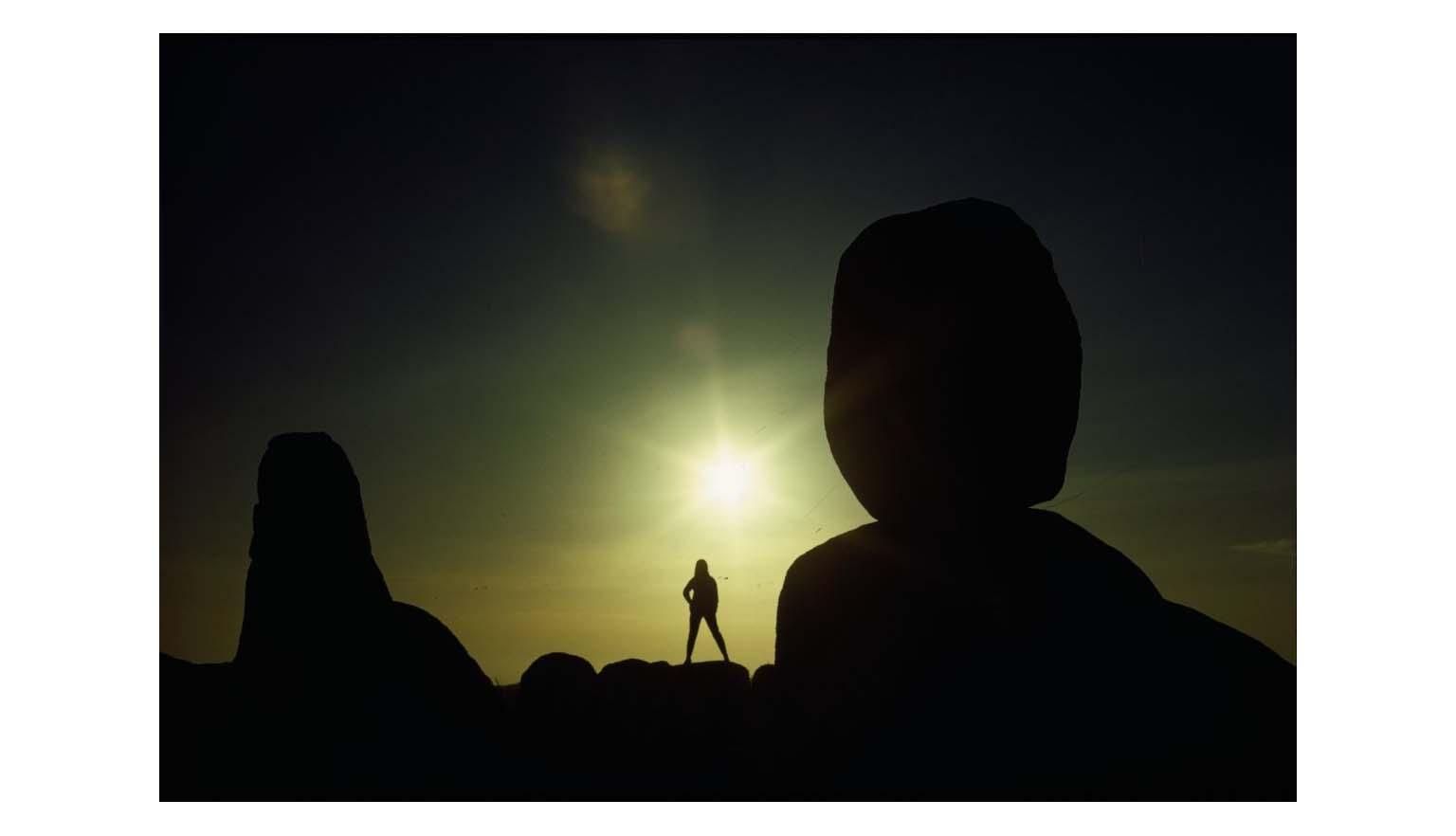 A high contrast image of two large monoliths silhouetted against a rising sun. The sky is shades of black, grey and yellow. A lone figure of a woman is silhouetted against the sun light.
