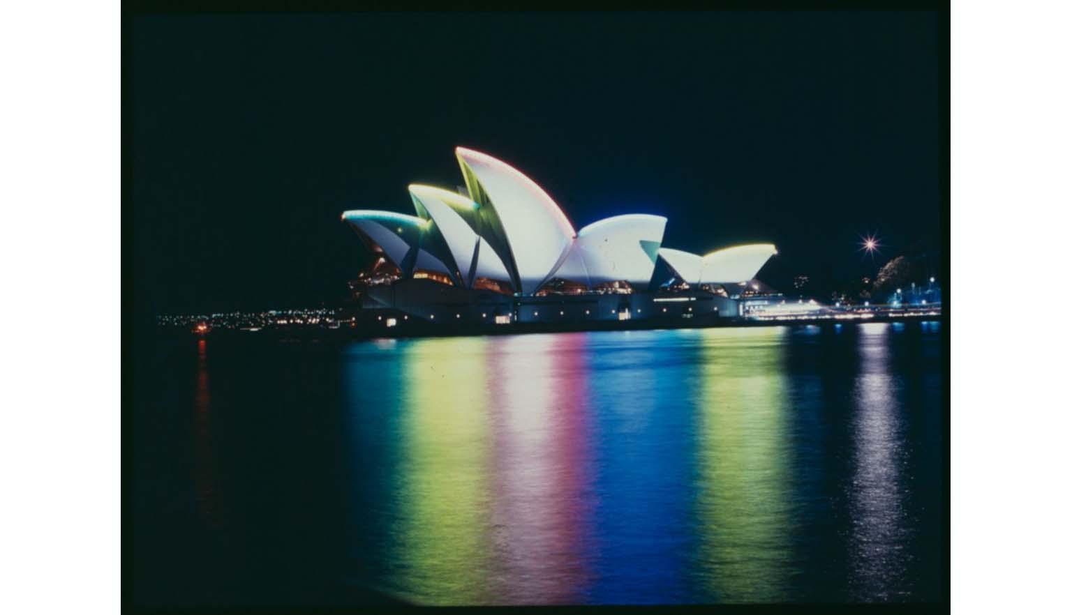 A nighttime photograph of the Sydney Opera House. The sails of the Opera House are lit up in different colours that are reflecting green, yellow, pink, blue and chartreuse into the water of the harbour.