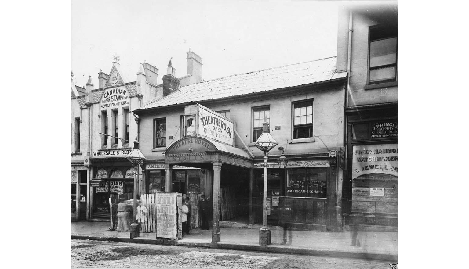 Black and white photograph of a row of shopfronts. One facade reads 'Theatre Royal: Open Every Evening'.