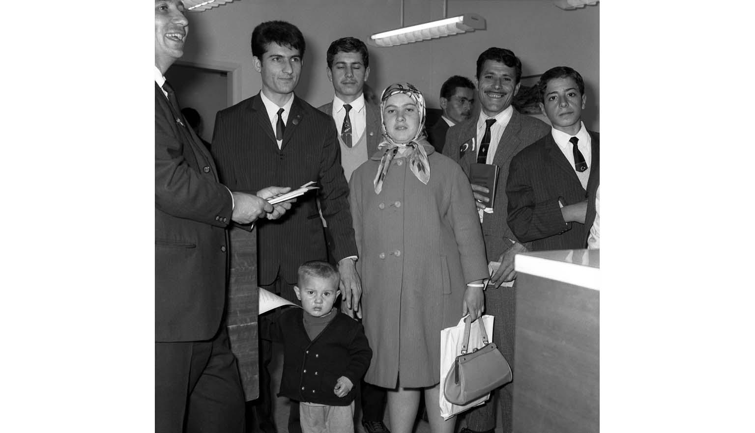 Eight people stand infront of a desk. There are 6 men, one woman and a small boy. The woman is wearing a headscarf and carrying a shopping back and a handbag. One man is holding tickets/papers. Another man holds a passport.