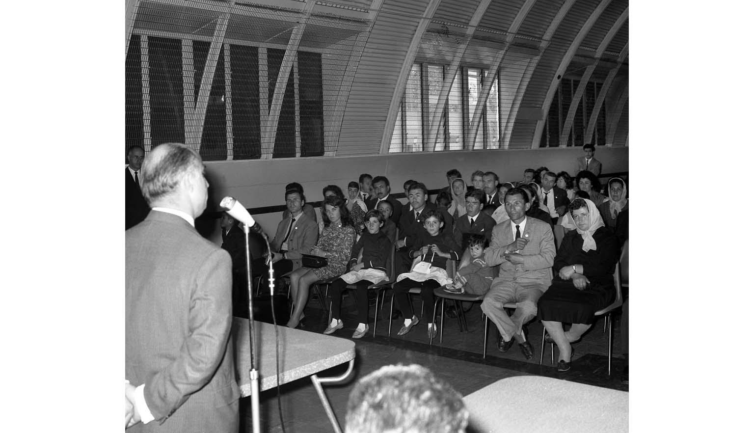 A photograph of a large group of people seated in an auditorium. A balding man is standing at a microphone addressing the audience.