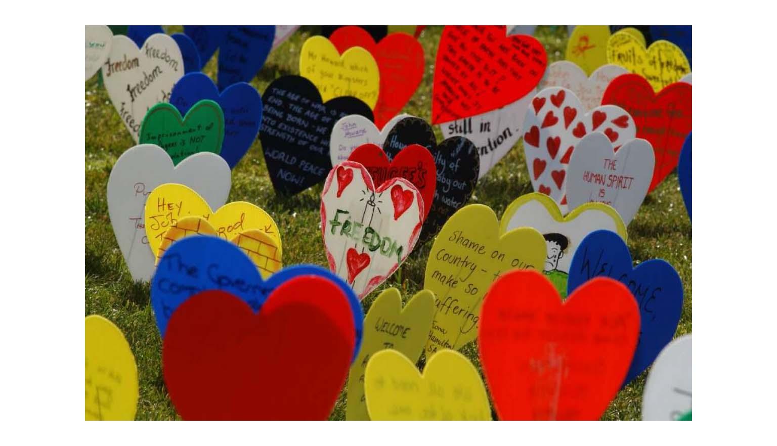 A photo of lots of different coloured plastic love hearts on a wire, stuck in a grassy area. The hearts are decorated with writing about freedom, and drawings.