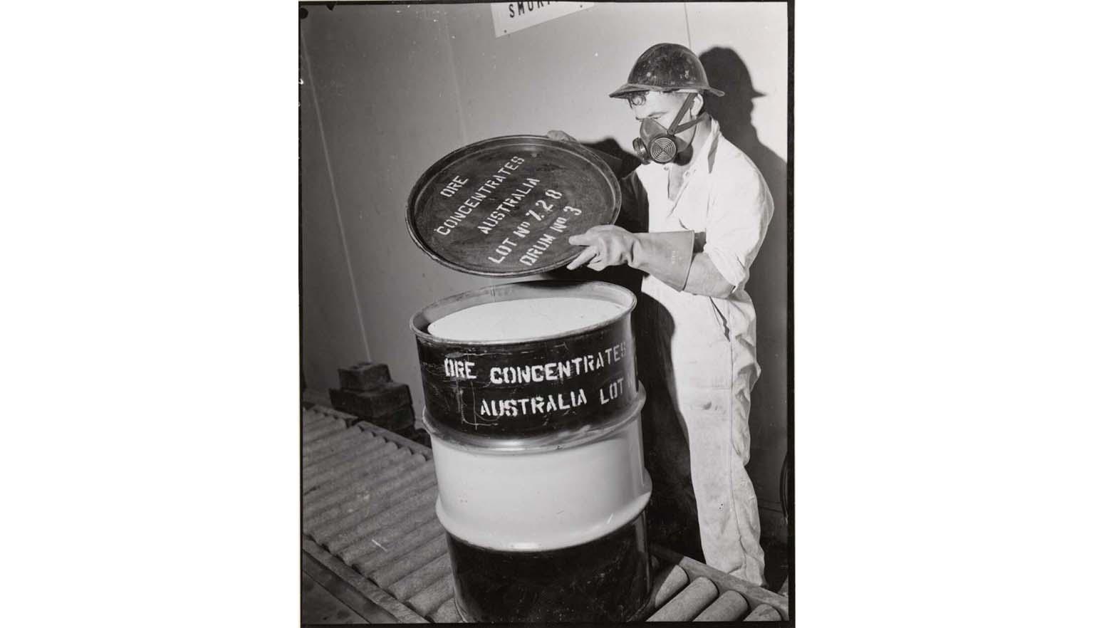 black and white photograph of a man in protective gear and a gas mask placing the lid on a barrel of uranium