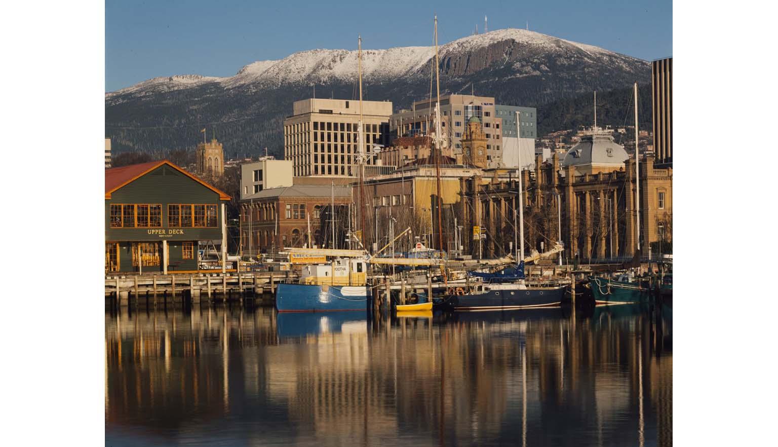 A city harbour scene. In the background a large snow capped mountain. There are several small boats on the water.