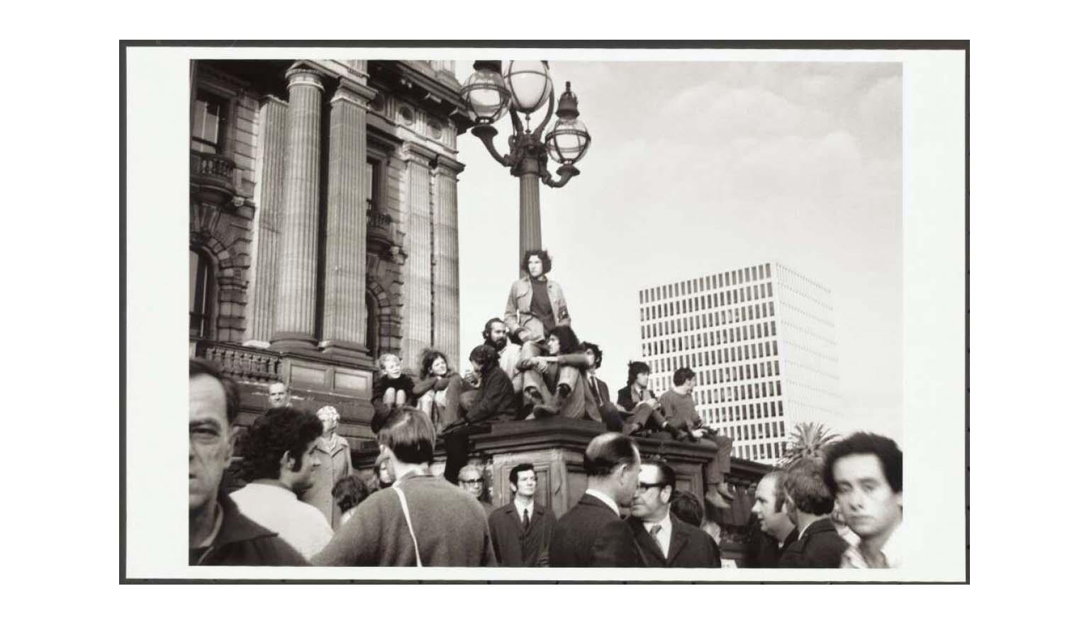 A black and white photo of tens of people gathered on top of and around a large light  on a large stone plinth in front of Victoria's Parliament House.