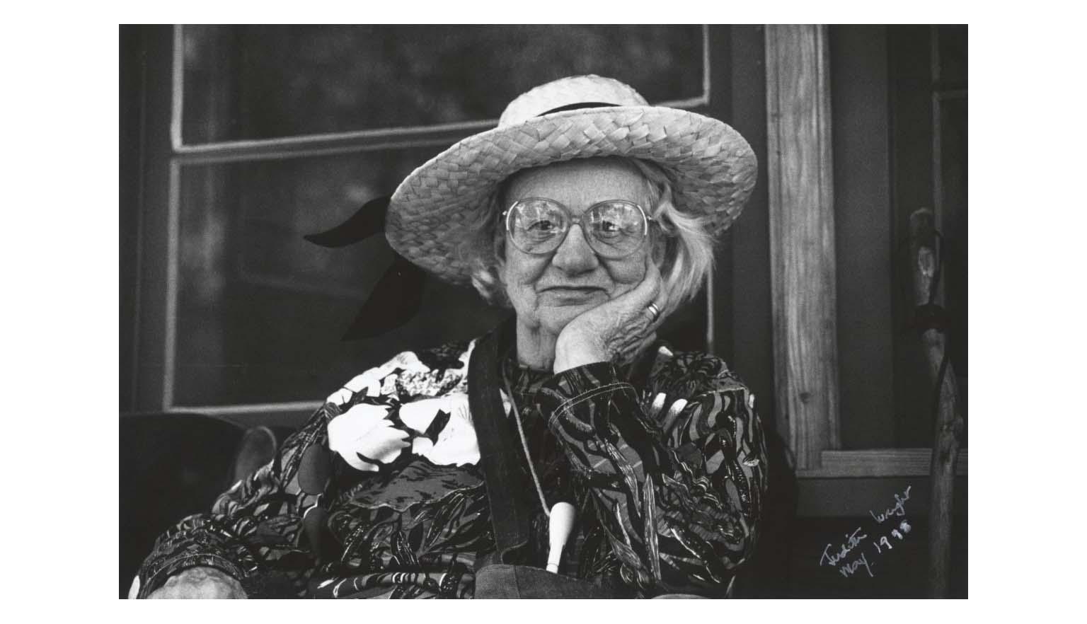 A black and white portrait photo of an old woman with white hair under a straw sunhat, wearing large reading glasses, and a long sleeved patterned dress, sits smiling at the camera with her hand resting, cupping the left side of her chin and face.