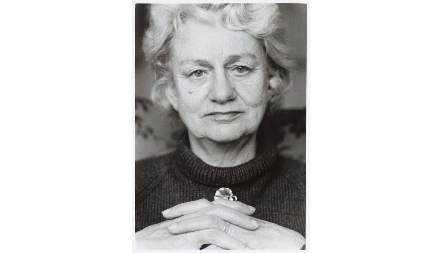 A black and white portrait photo of an elderly woman with short white, wavy hair wearing a black roll neck jumper and a small floral brooch just below the collar, with her interlaced fingers resting in the foreground.