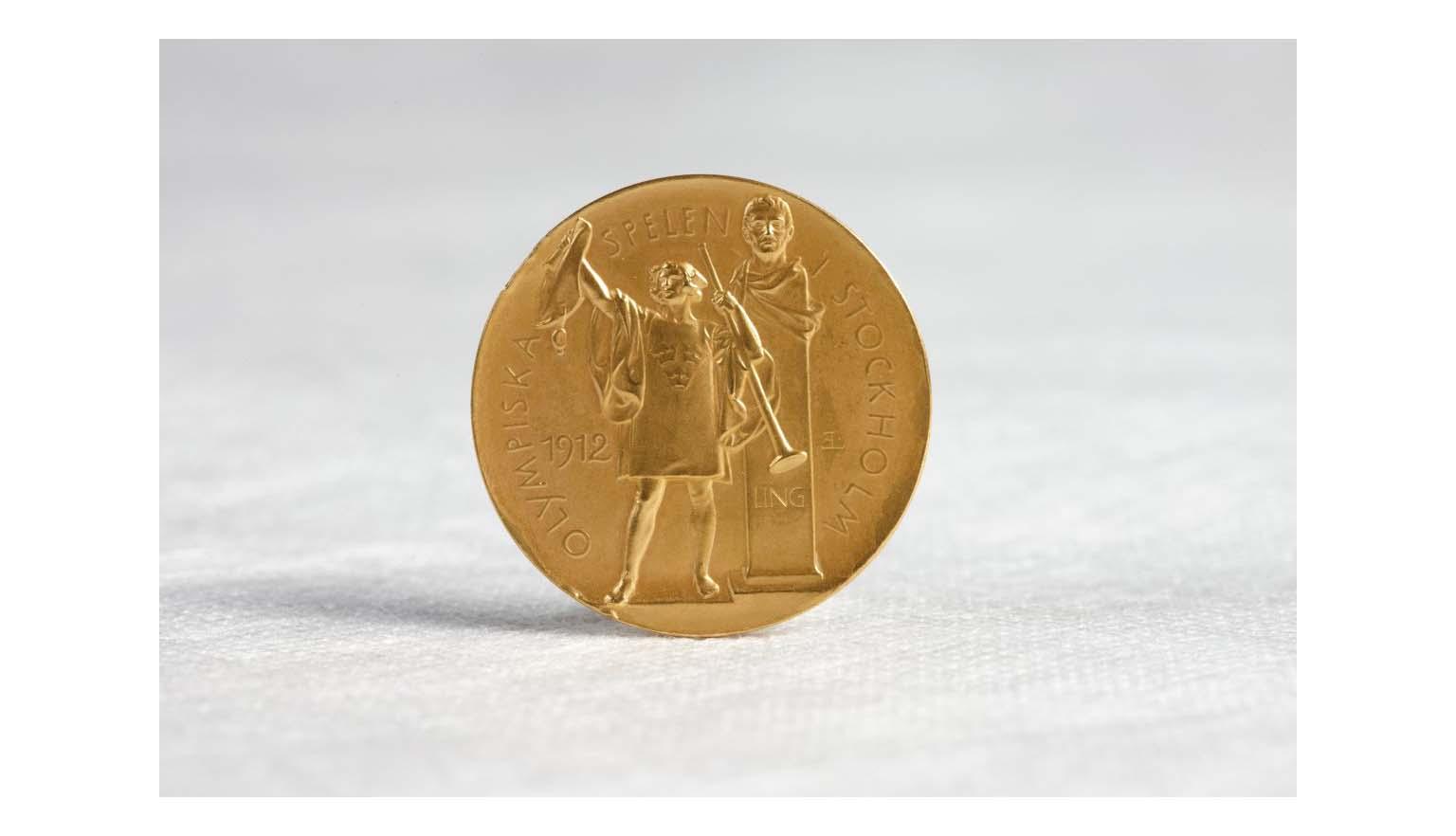 A gold medal standing upright on a white cotton surface. The medal shows a figure holding a trumped and a wine amphorae. Behind the figure is a bust on a stand. In raised lettering around the edge of the medal are the words 'Olympiska Spelen Stockholm' The year '1912' is also present.