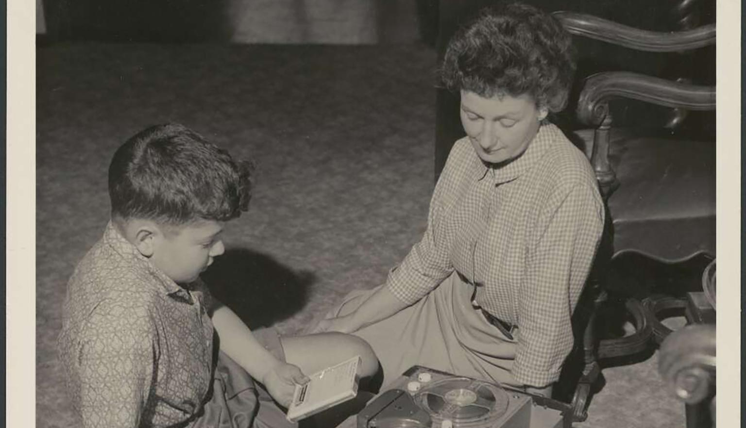 A woman sitting on the floor with a little boy listening to a reel to reel