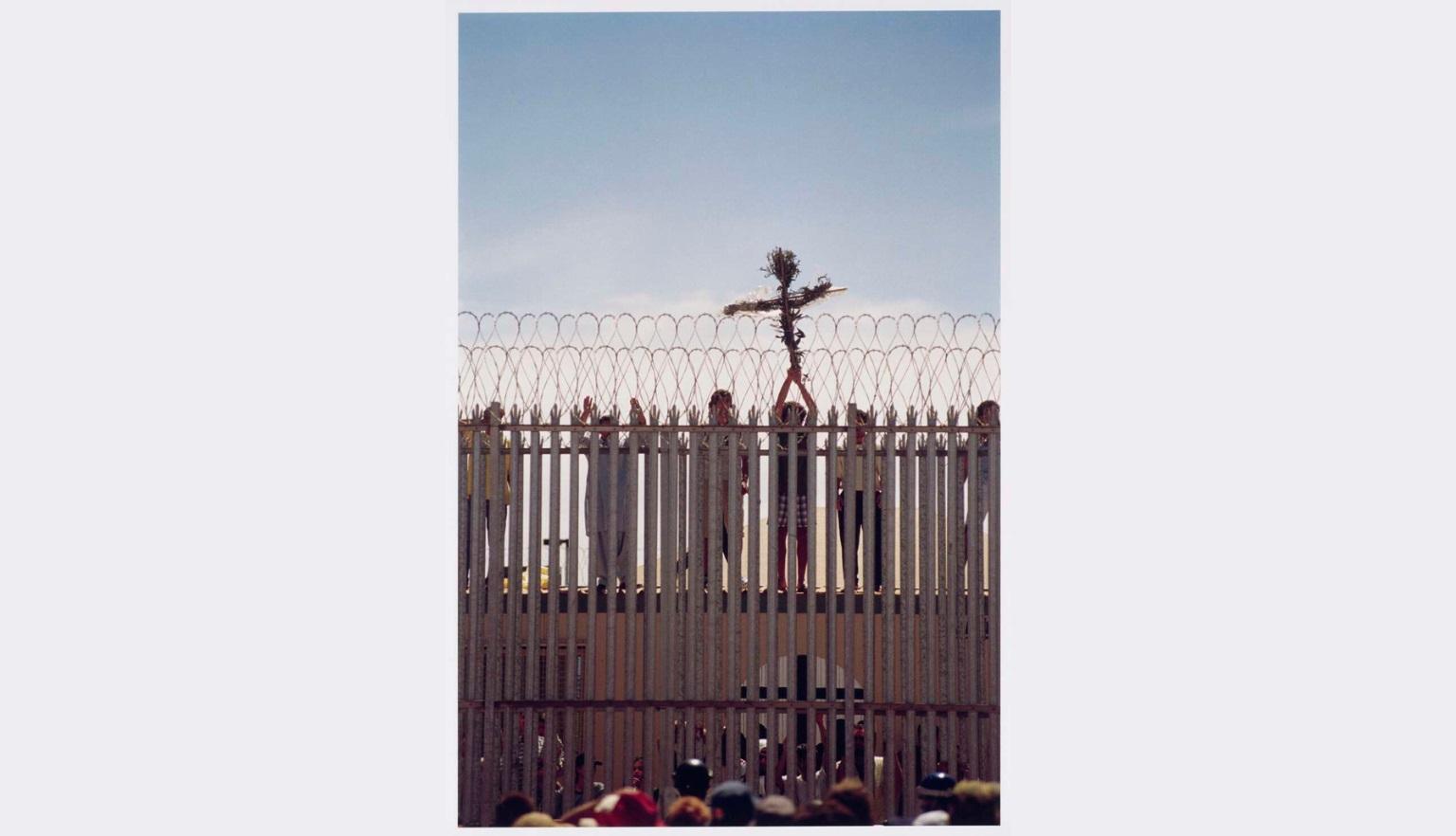 A wooden cross wrapped with foliage is held high by hands reaching through vertical metal bars topped with barbed wire.