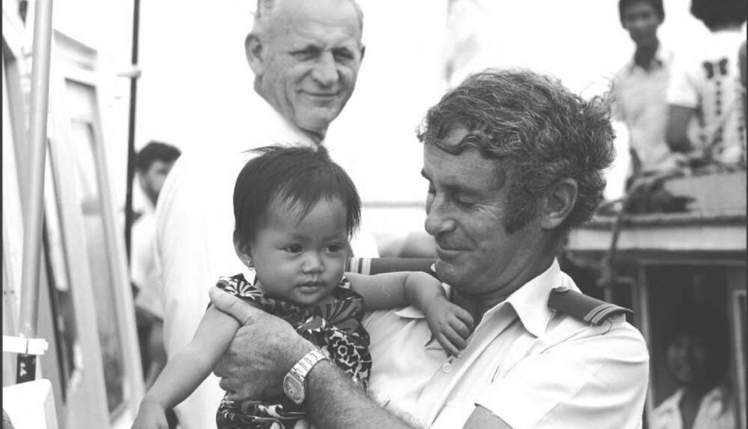 A customs officer cradles a young Vietnamese child while smiling warmly. Another man stands in the background on a boat surrounded by several people.