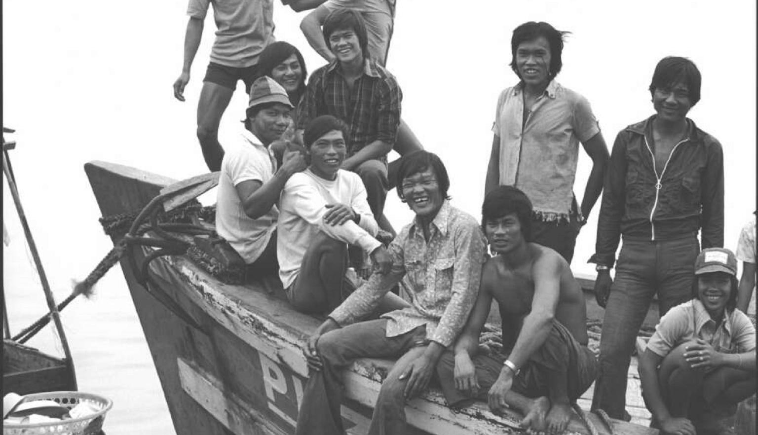 A group of smiling men sit and stand on the deck of a wooden boat, exuding camaraderie. 