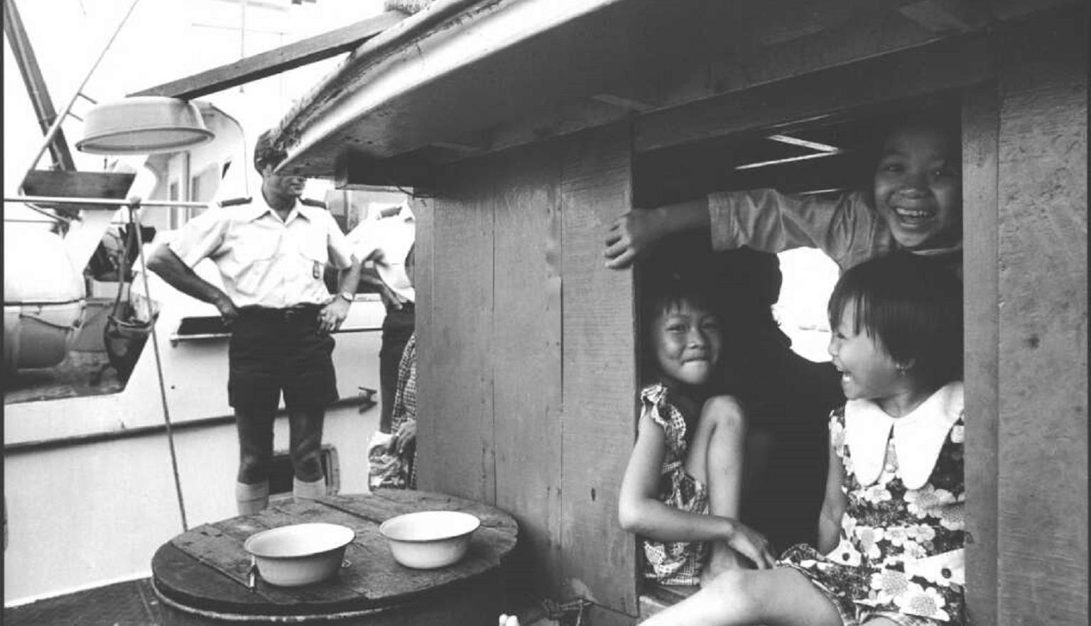 Three young girls smile and sit together inside a wooden boat cabin. A customs officer stands outside near a vessel labeled "Customs." Two bowls rest on a barrel near the cabin.