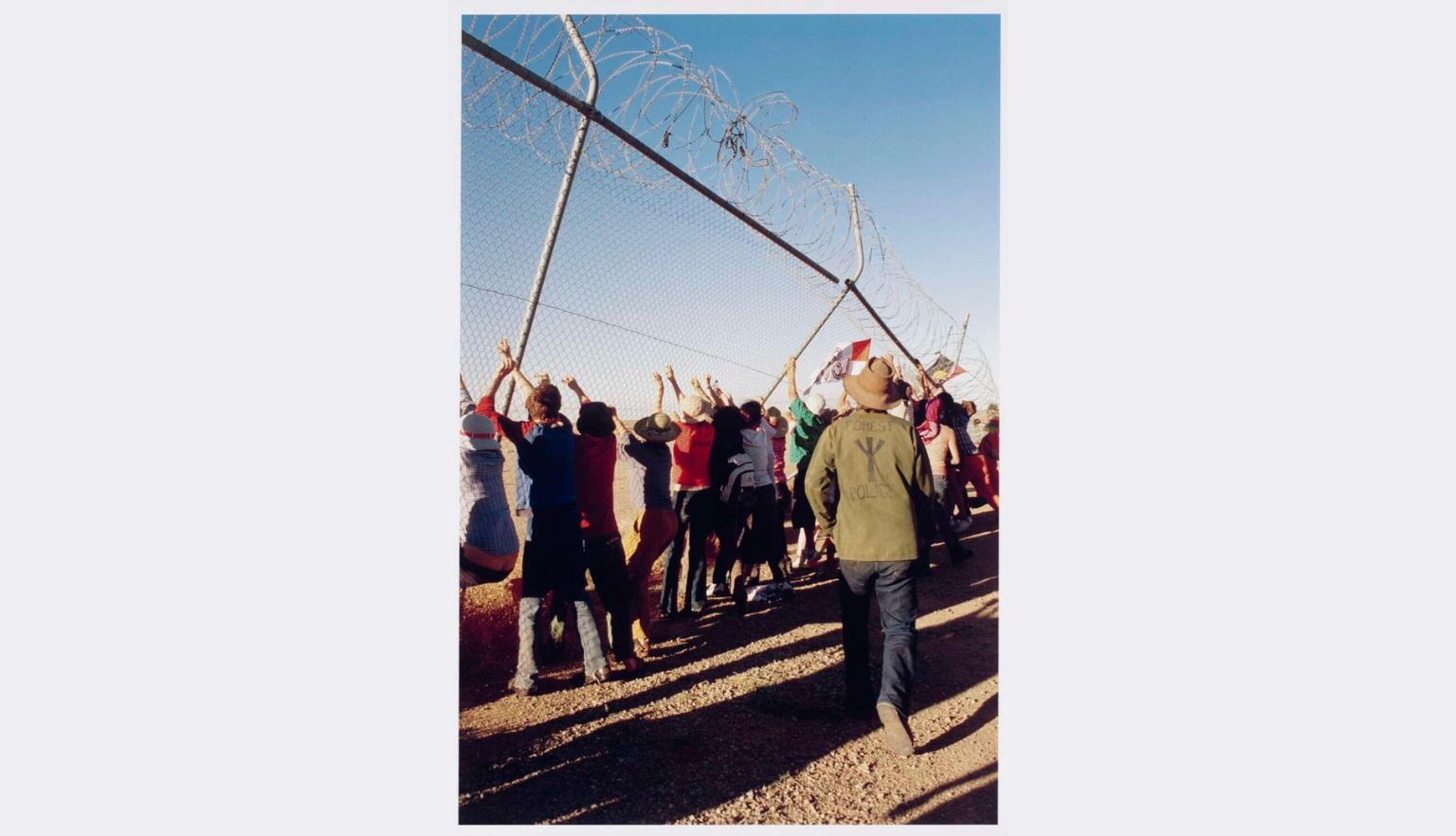 A group of people pull down a chain-link fence topped with barbed wire, their arms raised as the structure bends under pressure.