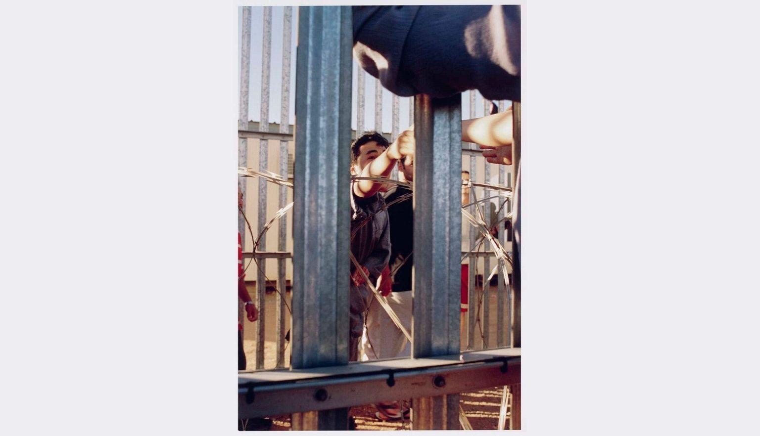 Hands of protestors stretch through metal bars to meet those of a person inside, with razor wire visible in the foreground