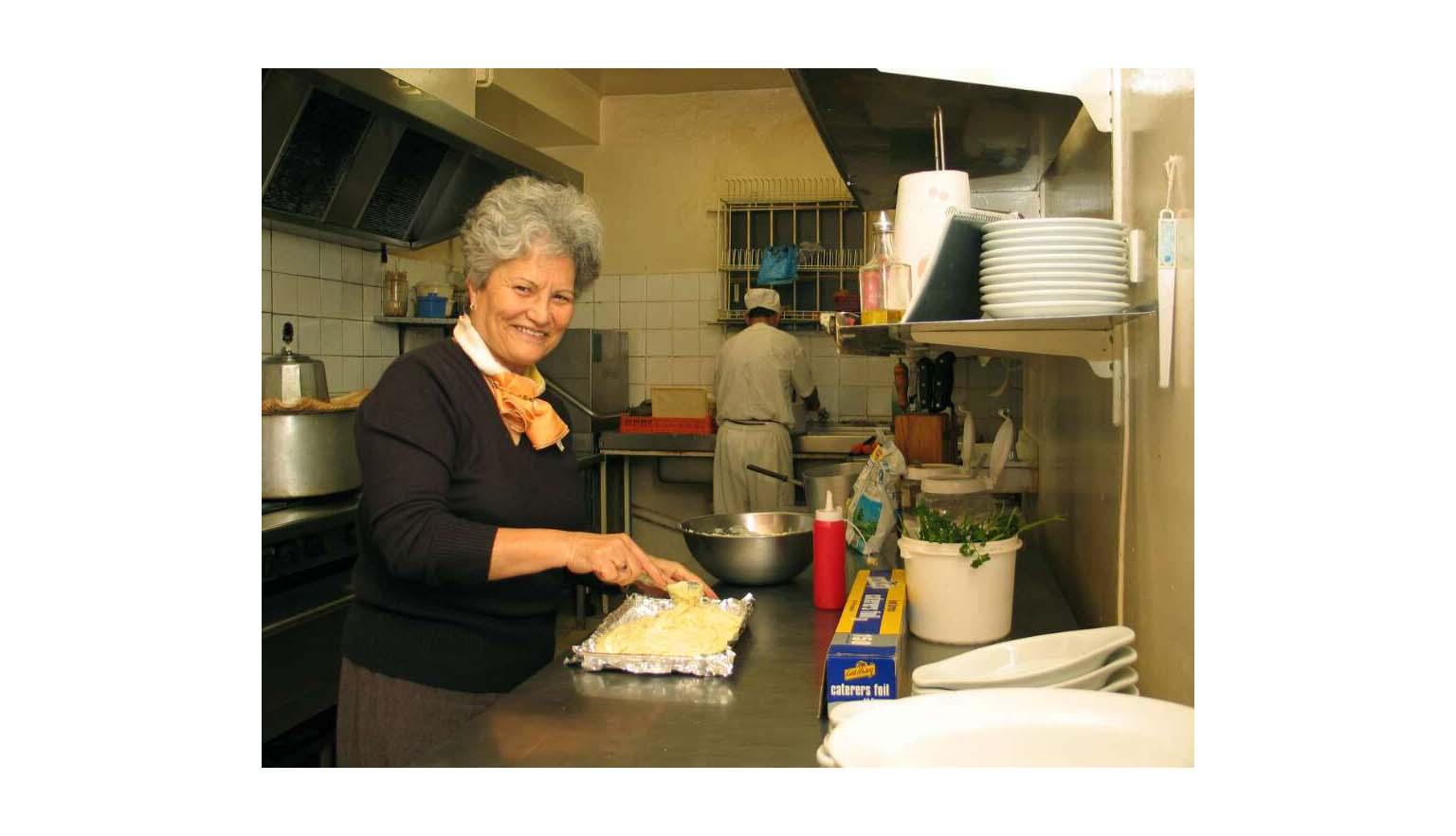 Woman with short grey hair smiling at the camera as she spreads food in a foil-lined tray in the kitchen of a restaurant
