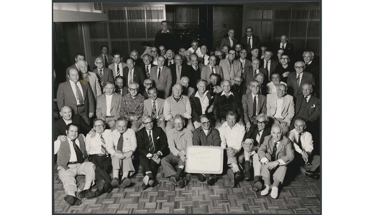 A black and white photograph of a large group of men, mainly older, posed for a group photo. Some men are sitting in the front rows, others stand at the back. They are all smiling at the camera. A man in the centre of the from row hold a white sign with black writing. It reads 'Dunera Reunion 1981'