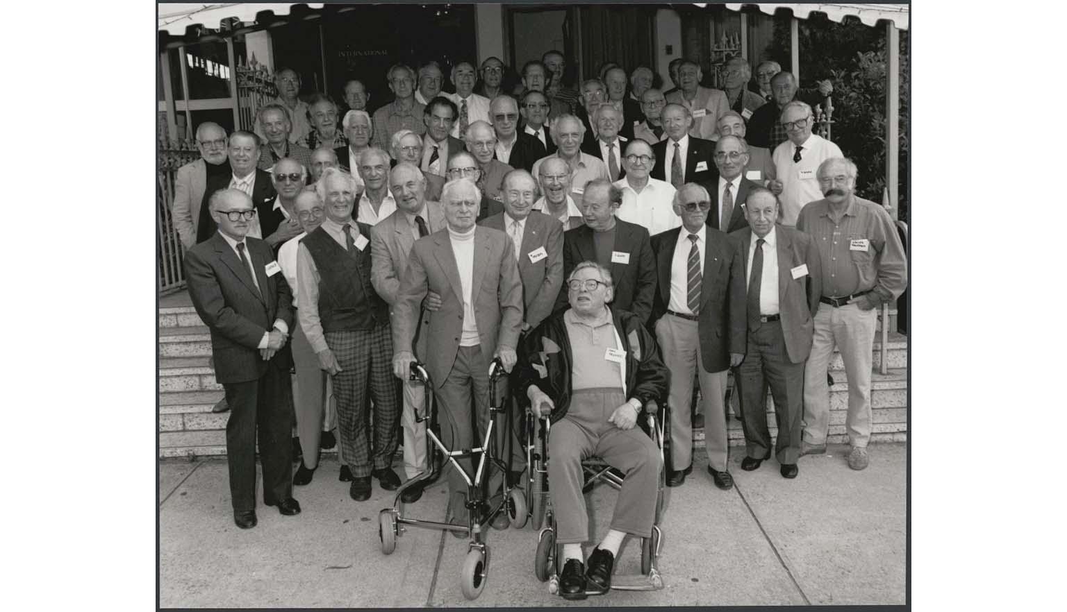 A black and white photograph of a large group of older men, posed for a group photo.  Most of the men are smiling at the camera.In the middle of the front row is a man sitting in a wheelchair. To his left a man stands holding a walking aide while a man holds his arm.