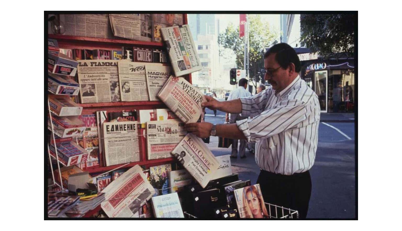 Man in striped button up shirt tucked into dark pants looking at newspapers in various languages at a kiosk on the street