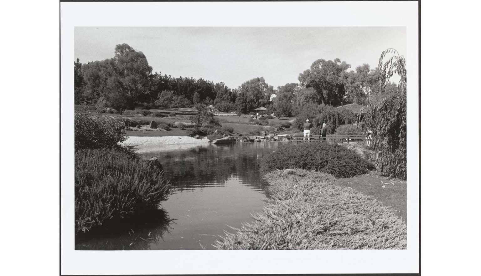 A black and white photograph of a manicured Japanese garden. There is a large calm lake in the centre of the image. There are large 